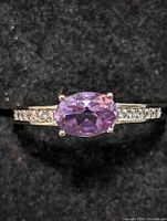 Close-up front view of oval purple amethyst gemstone set in sterling silver ring with small clear accent stones on band against black textured background