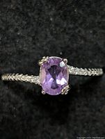 Close-up view of sterling silver ring with oval faceted purple amethyst gemstone and accent stones along the band on black background.