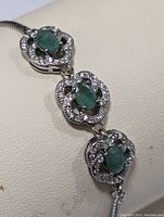 Close-up of silver bracelet showing three oval emeralds each surrounded by small clear stones within an ornate silver setting.