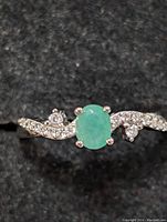 Close-up photo of sterling silver ring with oval emerald center and twisted band with clear accent stones on black background