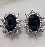 Pair of sterling silver earrings with oval sapphire center stones surrounded by clear cubic zirconia stones in a floral halo setting.