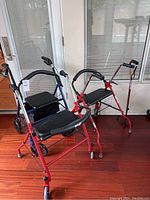 Three walkers and one cane arranged on a wooden floor near glass doors, showing overall view of lot items and condition.