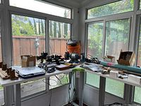 Two white adjustable-height tables forming an L-shape in a sunroom, loaded with rod-building tools and supplies.