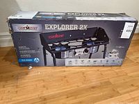 Box of Camp Chef Explorer 2X stove placed on wooden floor, showing product image and model name