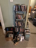 Metal rack loaded with jewel case music CDs and DVDs with some loose and out of cases on floor and in boxes nearby.