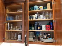 Photo showing two wooden kitchen cabinets open with shelves filled with various kitchen glassware, mugs, and plates.