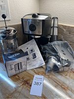 Full view of DeLonghi espresso machine on counter with French press, coffee grinder box, instruction sheet, and extra coffee accessories placed around.