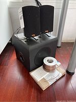 Two black Klipsch satellite speakers resting on top of the black subwoofer, all set on a wooden floor with coiled speaker wire and box.