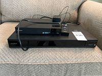Two Sony Blu-ray players stacked, one with remote and power adapter on top, placed on fabric couch.