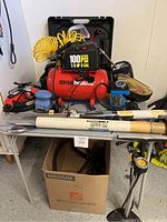 Lot arranged on white folding table showing Central Pneumatic red air compressor, Black and Decker electric sander, metal rods, blue hand sander, jumper cables, pressure gauge and coiled hose.