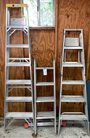 Three aluminum ladders next to a wood-paneled wall: two ladder frames and one smaller ladder between them.