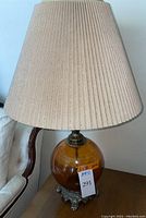 Full view of the amber glass globe table lamp with beige pleated shade and ornate metal base placed on a wooden surface next to a sofa.