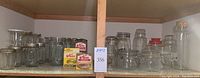 Wide assortment of clear glass mason jars and storage jars displayed on wooden shelves, some with lids and boxes of caps.