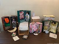 Five porcelain dolls in their own individual travel cases or trunks placed on a table, alongside certificates and small boxes for accessories.