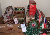 Wide shot showing multiple boxes filled with vintage Shiny Brite glass ball ornaments in various colors, boxed Lighted Musical Bell Cluster, boxed mini lights, tinsel, and other ornaments.