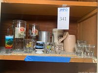 Shelf with multiple mugs and shot glasses including Dorothy Kindell diving woman mug, pair of San Francisco 49ers beer mugs, aluminum Playboy mug with clear base, and various shot glasses with prints and logos.