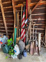 Wide view of all garden tools and accessories grouped against a wooden shed wall, including shovels, rakes, watering cans, blower, and more.