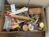 Box filled with various kitchen utensils such as rolling pin, graters, whisk, scissors, and other kitchen gadgets.