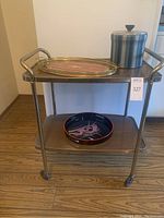 Bar cart with two wood veneer shelves and metal frame, holding the trays and ice bucket on top and bottom shelves.