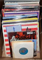 Wide view of cardboard box filled with assorted vinyl LP album covers and 45 RPM singles, showing colorful album artwork and visible wear.