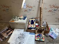 Wide shot showing white woven sewing basket, small sewing boxes with thread, yardsticks, jars and boxes with buttons, lace trims, craft paint bottles, scissors, and sewing patterns.