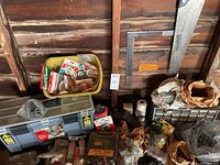 Photo showing grey plastic tool box with lid closed, various hardware packages, hand trowels, miscellaneous parts, saw, and metal framing square against wood paneled wall.