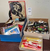 Three boxes filled with assorted travel souvenirs including postcards, swizzle sticks, collector spoons, keychains, and décor items.