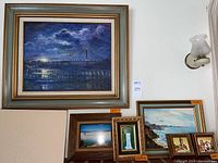 Large framed oil painting showing the Golden Gate Bridge at night with reflections on water and dramatic sky, accompanied by smaller framed paintings and a framed photo, all arranged on a wall.