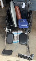 Photo showing black Drive wheelchair with extended metal footrests, brown leather lumbar support pillow on seat, blue Medtronic Carelink Monitor case, and wheelchair seat cushion.