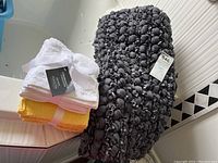 Photo showing tufted dark grey fabric bath mat with knotted texture and two bundles of terry facecloths in white and yellow tied with white ribbons and branded tags.