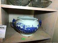 Blue Willow ceramic pot with scenic blue pattern, scalloped rim, and two blue handles placed on a wooden shelf.