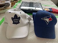 Photo showing both caps, Toronto Maple Leafs cap on left in white and Toronto Blue Jays cap on right in blue.