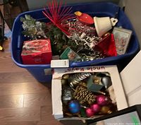 Blue tote filled with Christmas lights, red decorative piece, white ceramic bell, coaster set, pine garland, and a Mikasa bowl visible on floor.