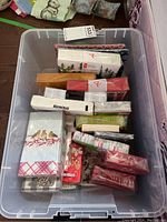 Top down view of the clear plastic bin containing multiple packages of Christmas-themed paper napkins and packs of paper plates, showing assorted colors and patterns.
