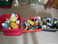 Three plastic bins filled with various cleaning products including fabric softeners, sprays, and gloves on carpeted floor.