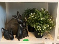 Shelving display of two bunny figurines, faux greenery plants in black pots, and a brown decorative vase.