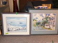 Two framed prints side by side on a surface, one depicting a coastal scene by C. Holding and the other a village street scene by Elizabeth Berry.