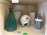 Photo showing full set of vases and pots on shelf: green textured glass vase, white ceramic pot, round tripod decorative vase, cylindrical off-white vase, and blue/white striped vase.