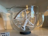 Clear glass KitchenAid mixing bowl with white beaters inside, showing measurement markings and the KitchenAid logo.
