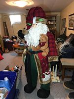 Full view of life size Santa Claus doll standing in a room with basement setting, showing outfit and attached teddy bear.