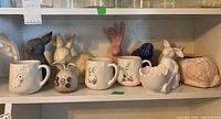 Shelf with white ceramic mugs with bunny illustrations and various bunny figurines in different colors and shapes