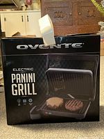 Front view of Ovente labeled box showing electric Panini grill with sandwich grilling plate design and Ovente branding.