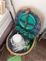 Top-down view of open wicker picnic basket showing green plastic plates held by leather strap, four green plastic mugs secured by leather straps, paper plates, plastic cutlery, and green plastic cups inside.