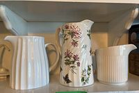 Three ceramic pitchers displayed on a shelf: one floral decorated, one white ribbed, one white fluted.