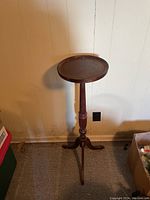 Front view of the wooden plant stand showing circular top, turned central column, and tripod carved legs against a white wall and carpeted floor.