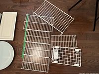 Seven white wire shelf organizer racks arranged on wooden floor, showing different sizes and styles including a rack with foldable legs.