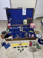 Overview of wooden jewelry box open displaying assorted costume jewelry including bracelets, necklaces, pendants, brooches, watches, and earrings on floor.