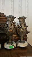 Front view of both metal cherub urns on marble bases with repair sticker