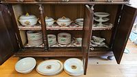 Multiple pieces of the French earthenware set displayed in an open wooden cabinet, showing plates, bowls, serving dishes, and tiered plates.