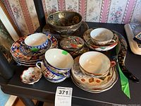 View of the grouped Asian-themed ceramic cups, saucers, plates, enameled bowl, and curved brush showing overall lot contents and condition